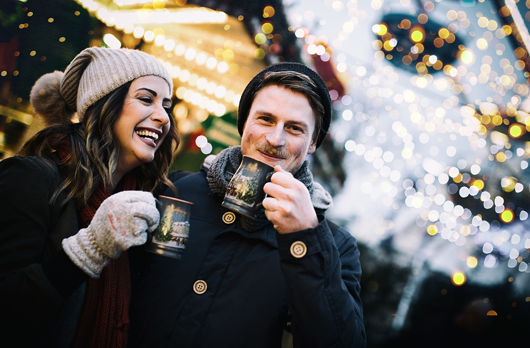 „Ein lachendes Paar trinkt heißen Punsch auf einem festlich beleuchteten Weihnachtsmarkt.“